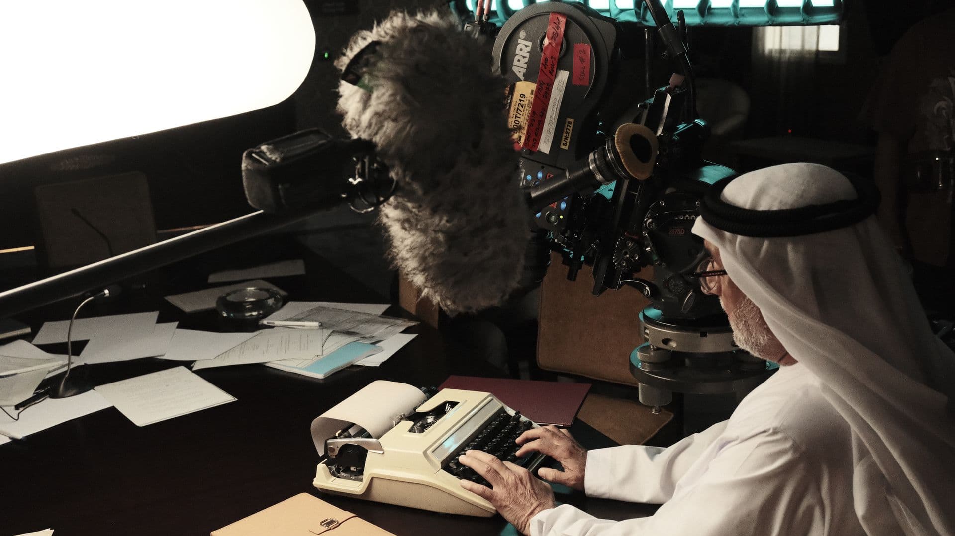 A man in a ghutra at a typewriter, a cinema camera in the foreground