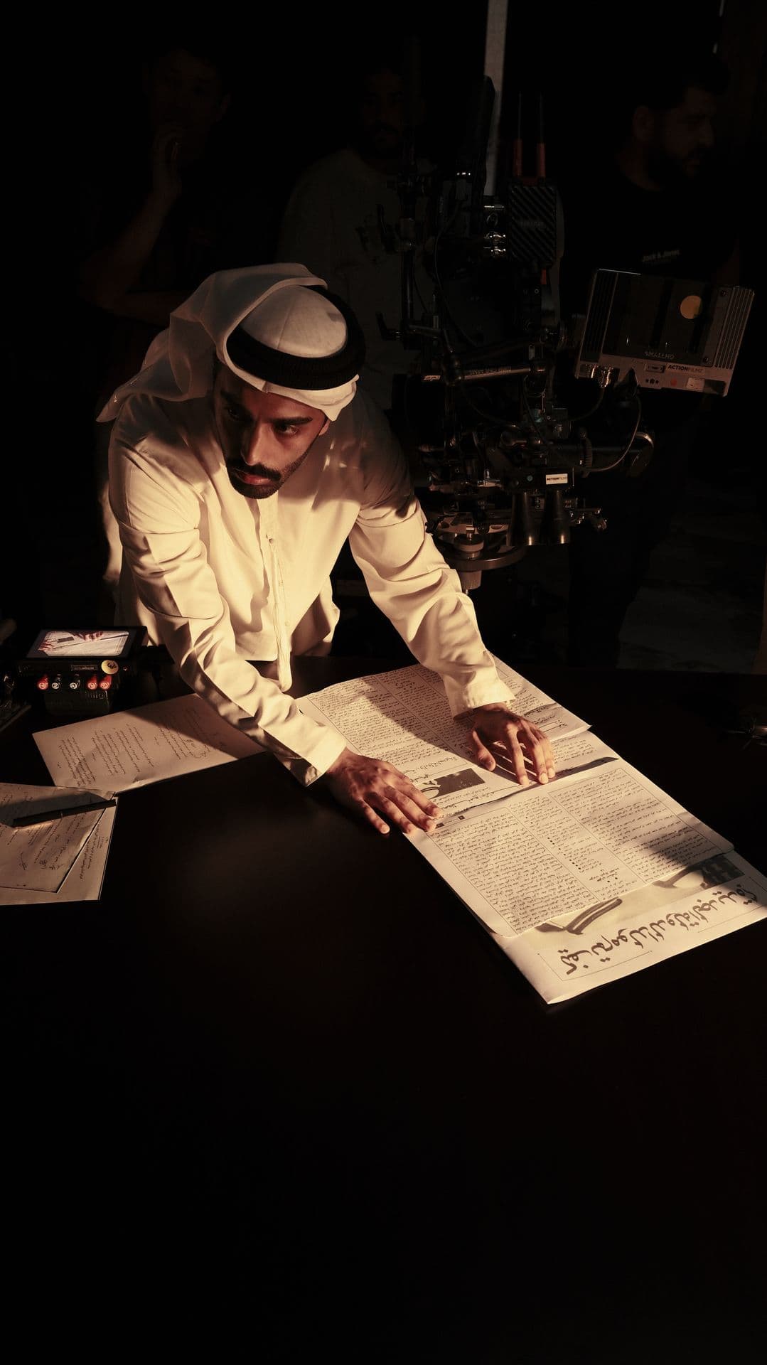 A man in a kandura writing at a desk under a single light